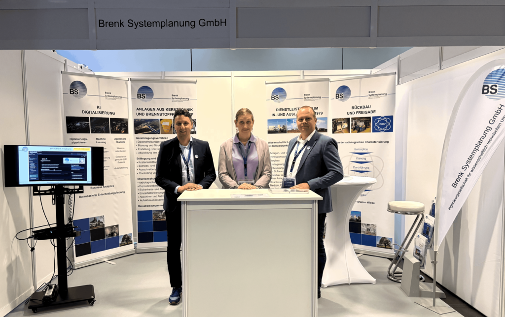 Drei Personen stehen hinter einem Messestand der Firma Brenk Systemplanung GmbH. Der Stand ist mit Informationsbannern ausgestattet. Links befindet sich ein Bildschirm mit einer Präsentation, rechts ein Stehtisch und ein Hocker. Die drei Personen tragen Business-Kleidung und blicken in die Kamera.