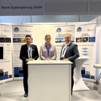 Three people are standing at an exhibition booth, with the sign “Brenk Systemplanung” above them.