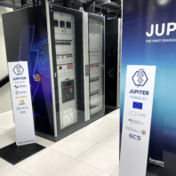 Rechenzentrumsbereich mit mehreren großen Server- und Schrankgestellen im Hintergrund sowie zwei aufgestellte Roll-Ups mit Aufschrift „JUPITER“ im Vordergrund; am Boden sind Kabelschächte und Fliesen zu sehen.