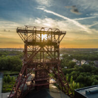 Ein Förderturm aus Stahl am Ufer eines Flusses, aus der Vogelperspektive bei Sonnenuntergang fotografiert; im Hintergrund liegt eine Stadt mit grünen Bäumen.