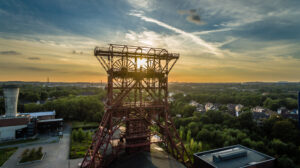 Ein Förderturm aus Stahl am Ufer eines Flusses, aus der Vogelperspektive bei Sonnenuntergang fotografiert; im Hintergrund liegt eine Stadt mit grünen Bäumen.