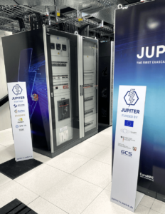 Rechenzentrumsbereich mit mehreren großen Server- und Schrankgestellen im Hintergrund sowie zwei aufgestellte Roll-Ups mit Aufschrift „JUPITER“ im Vordergrund; am Boden sind Kabelschächte und Fliesen zu sehen.