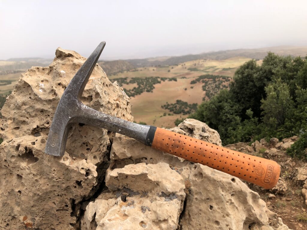 Geologischer Hammer mit orangefarbenem Griff liegt auf hellen, verwitterten Kalksteinen; im unscharfen Hintergrund eine weite, leicht hügelige Landschaft mit Feldern und vereinzelten Büschen unter hellem Himmel.