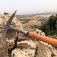 Ein Geologenhammer mit orangefarbenem Griff steckt in zerklüftetem Gestein; im Hintergrund sind Hügel und eine bewaldete Landschaft bei nebligem Himmel zu sehen.