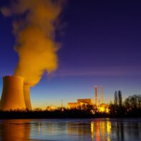 Ein Kernkraftwerk mit rauchenden Schornsteinen bei Sonnenuntergang, mit Spiegelung im ruhigen Fluss.
