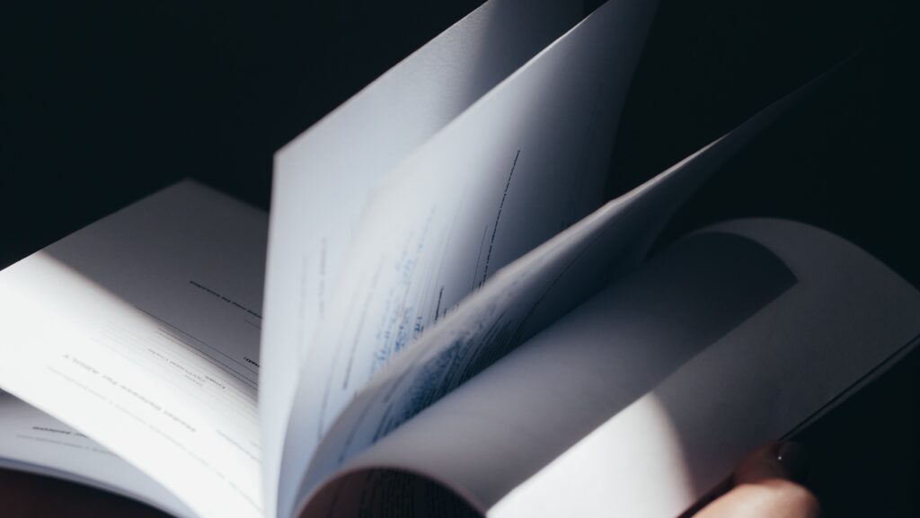 Blattseiten eines aufgeschlagenen Hefts oder Magazins, in sanftem Seitenwechsel: nahe Totale, weiße Seiten mit gedrucktem Text, einige Blätter sind im Flug; weiches Licht von links wirft Schatten und hebt die gebogenen Ränder hervor.