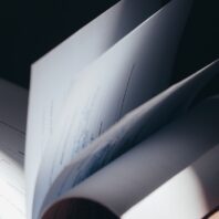 Nahaufnahme eines geöffneten Buchs mit gedrehten, unscharf wirkenden Seiten; im Vordergrund sind weitere Seiten im Schatten zu erkennen, das Bild wirkt dunkel und abstrakt.