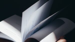 Nahaufnahme eines geöffneten Buchs mit gedrehten, unscharf wirkenden Seiten; im Vordergrund sind weitere Seiten im Schatten zu erkennen, das Bild wirkt dunkel und abstrakt.