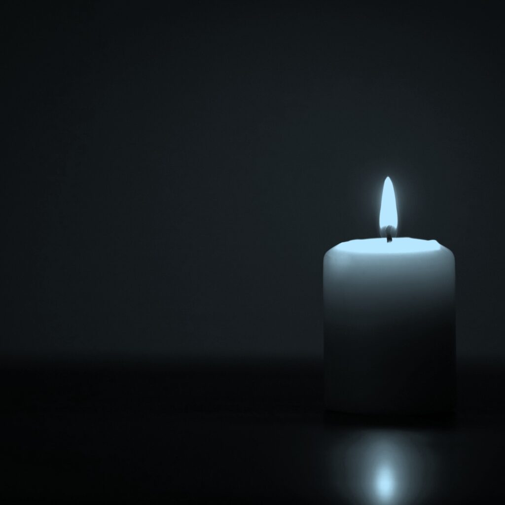 Eine weiße Kerze mit flackernder Flamme steht rechts im Bild vor dunklem Hintergrund, mit weicher Lichtreflexion auf dem Boden.