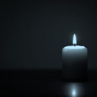 Eine weiße Kerze mit flackernder Flamme steht rechts im Bild vor dunklem Hintergrund, mit weicher Lichtreflexion auf dem Boden.
